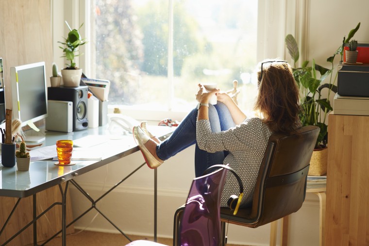 Image: Pensive woman looking through window with feet up on desk in sunny home office